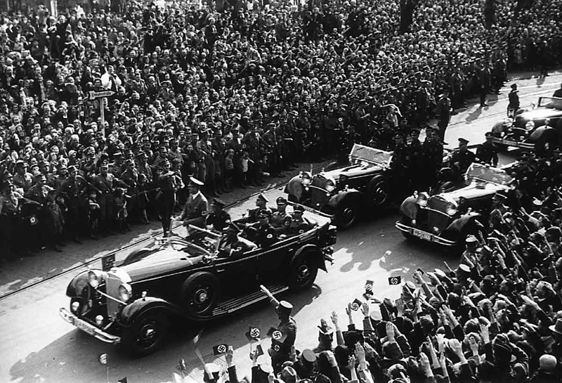 Adolf Hitler is standing in his open car while driving through Köln (Cologne)